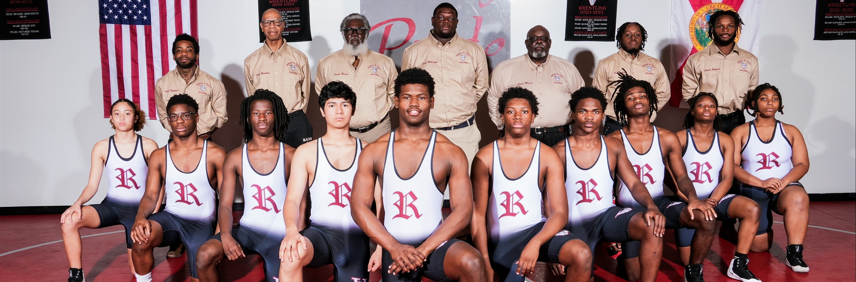 A wrestling team stands on a red mat, wearing white jerseys. Behind them, a white wall displays an American flag and other banners.