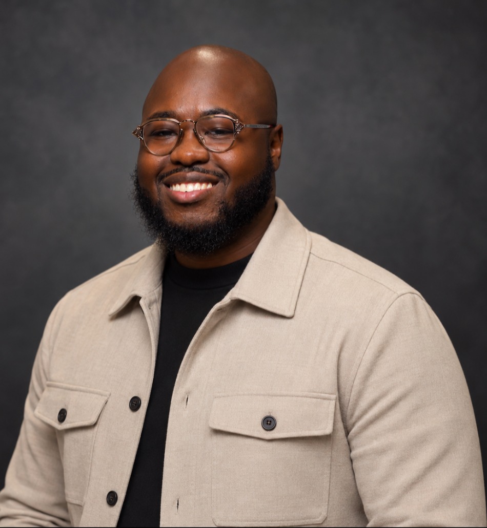 A smiling man with glasses, beard, and a beige jacket stands against a dark background.
