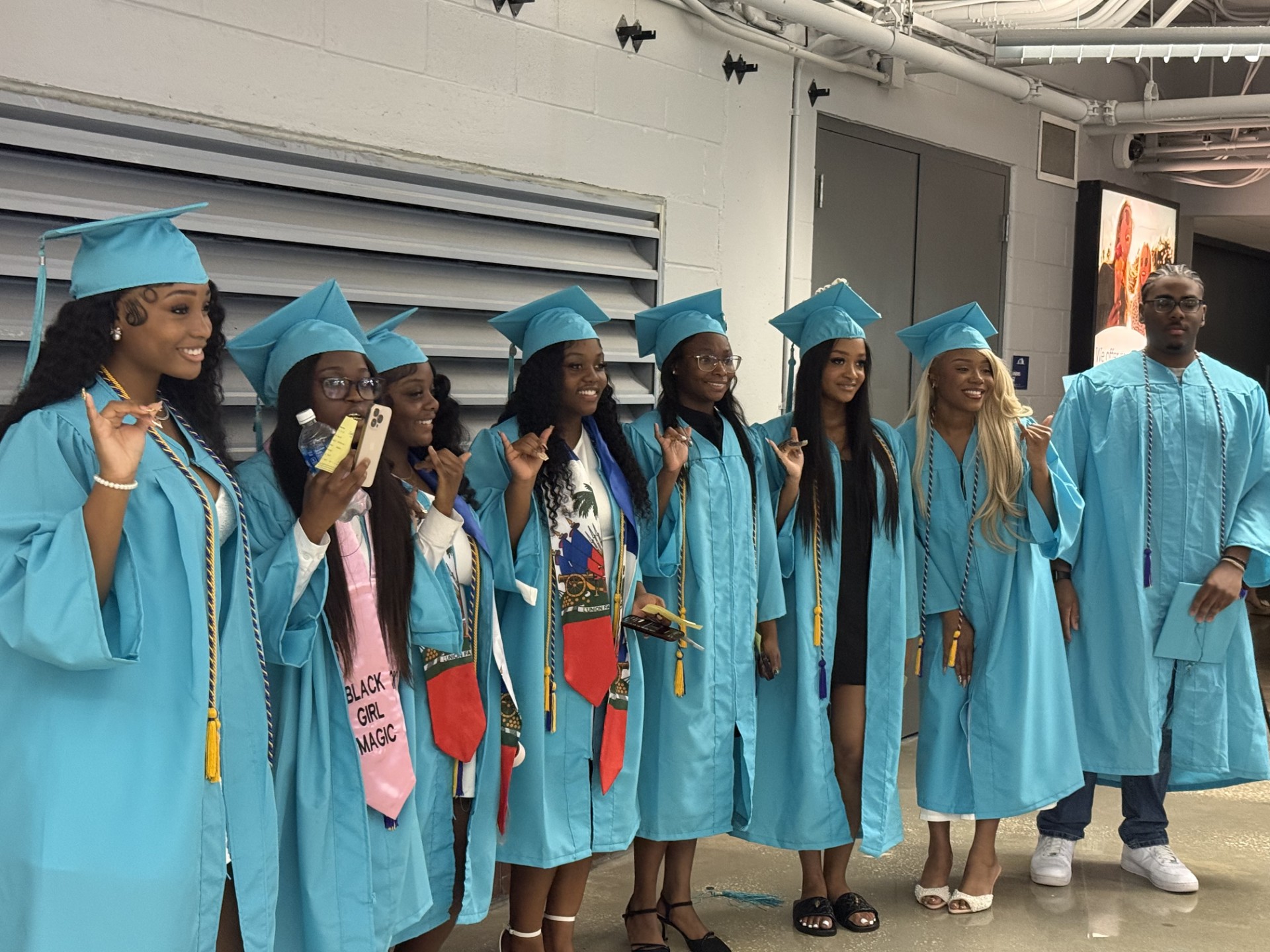 A group of graduates in blue gowns and caps, one holding a banner, pose in a hallway.