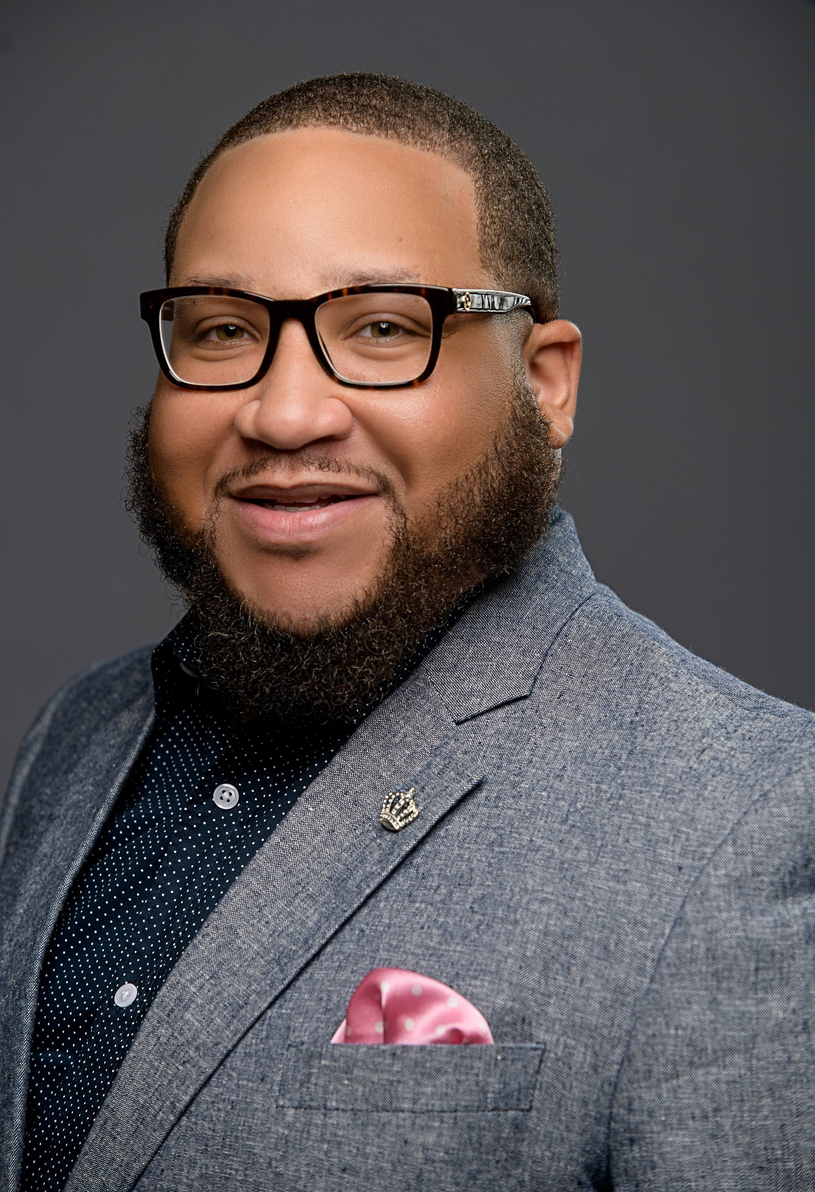 Man in gray suit with a black beard and glasses. He wears a blue shirt with a pink pocket square.