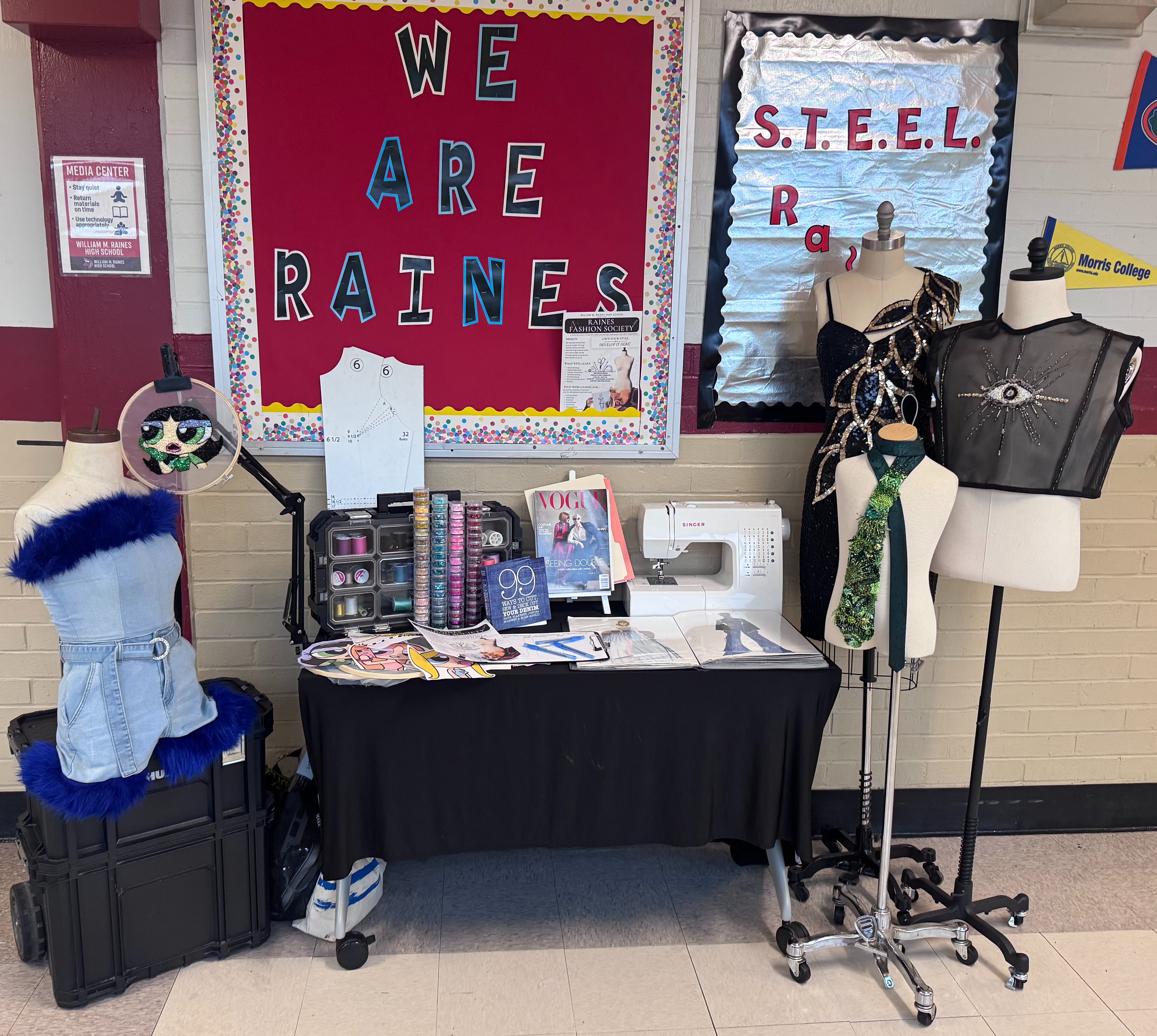 A table displays a sewing machine, books, and costumes with mannequins. Behind it, a bulletin board reads "We are Raines".