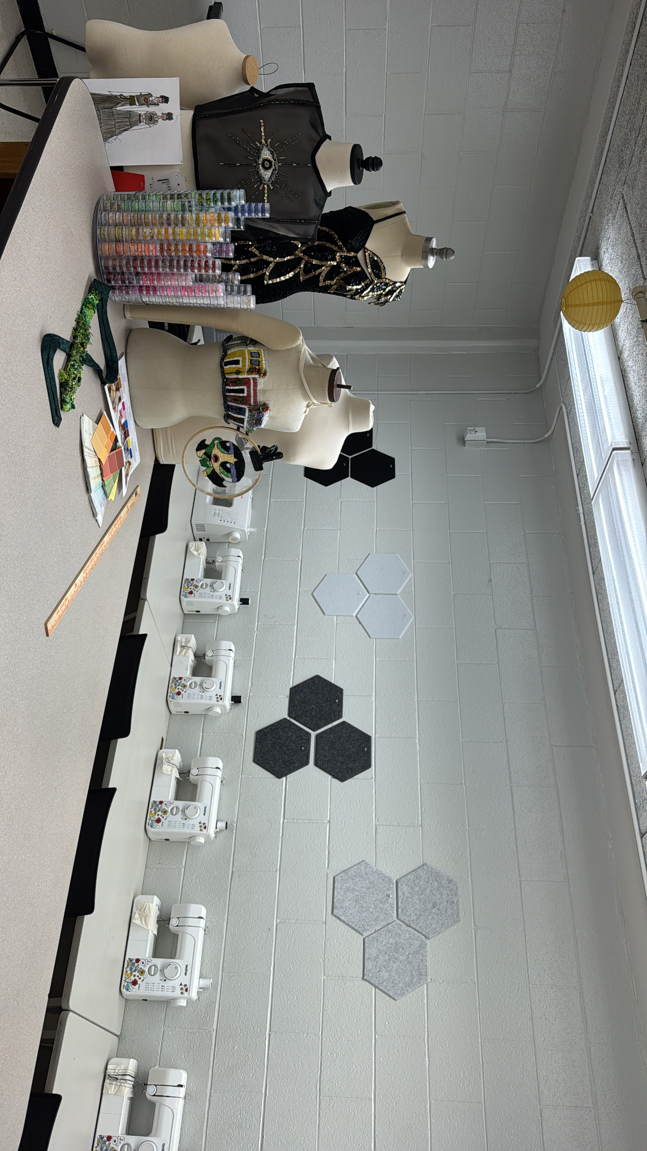 A room with white walls and floor displays mannequins, sewing machines, and a table with assorted fabrics and craft supplies.