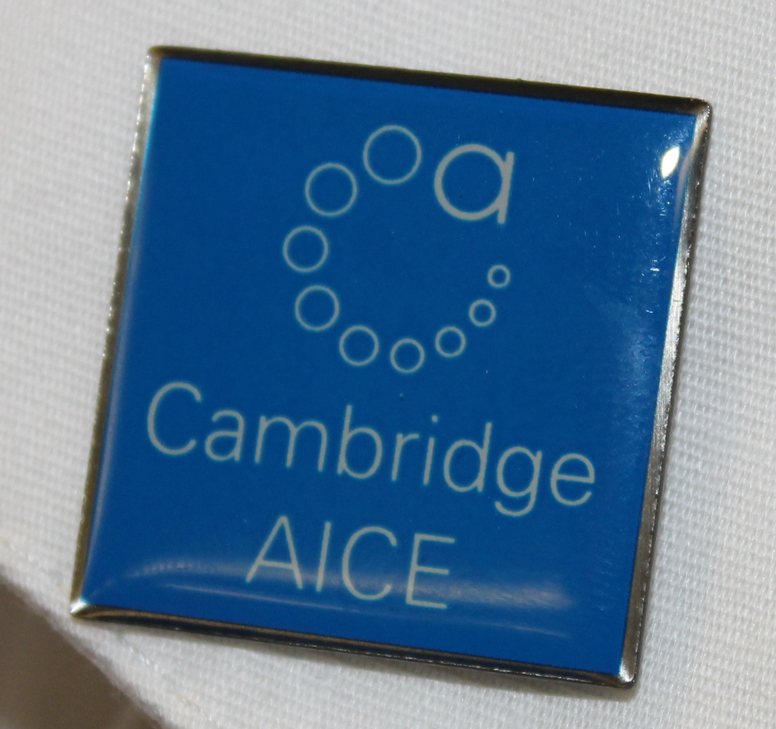 A blue pin with white text reading "Cambridge AICE" on a white shirt collar with a black background.