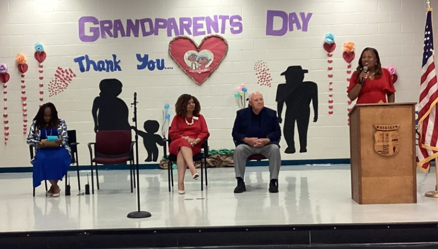 image of stage with a speaker at a podium and three adults sitting in chairs, "Thank You Grandparents" banner is behind them