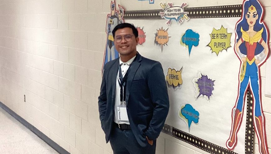 Image of a male teacher standing in a school hallway beside a bulletin board.