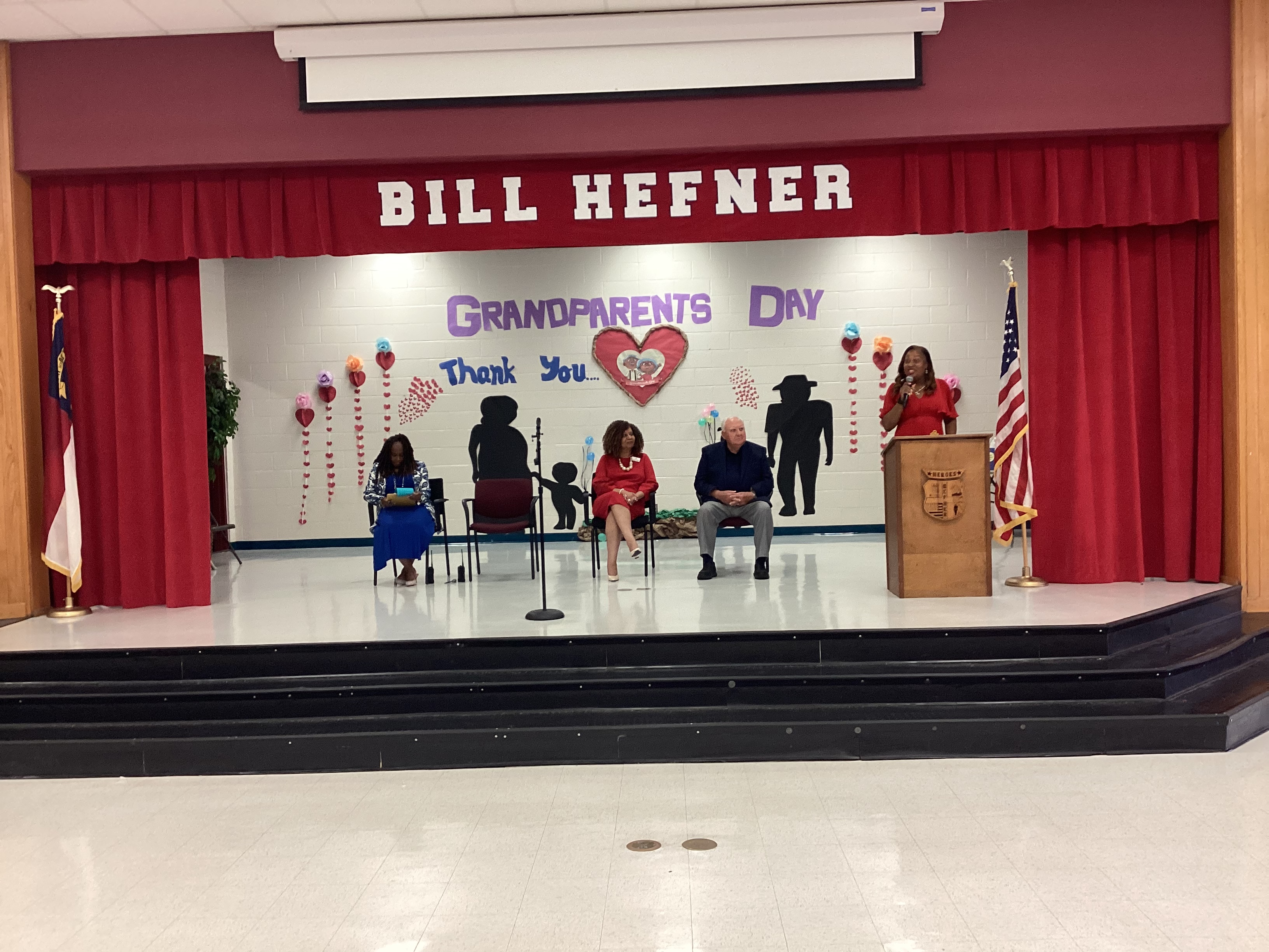 image of image of stage with a speaker at a podium and three adults sitting in chairs, "Thank You Grandparents" banner is behind them