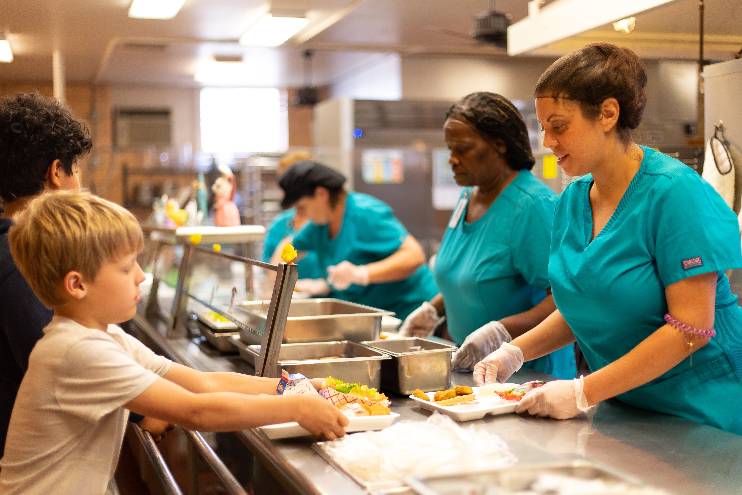 A photo of children going through the lunch line in their cafeteria, lunch ladies are serving.