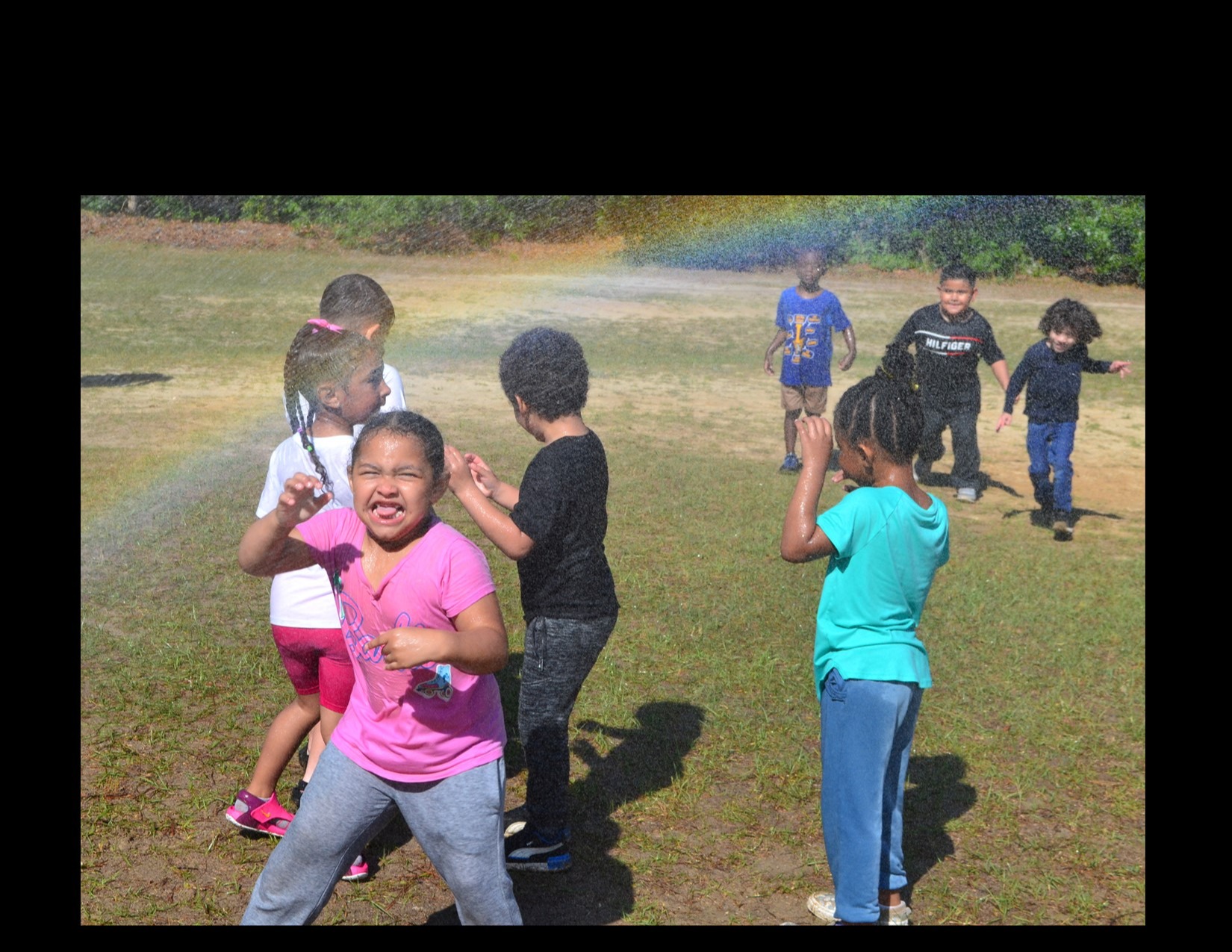 Several kindergarten children playing in teh water spray from a hose.