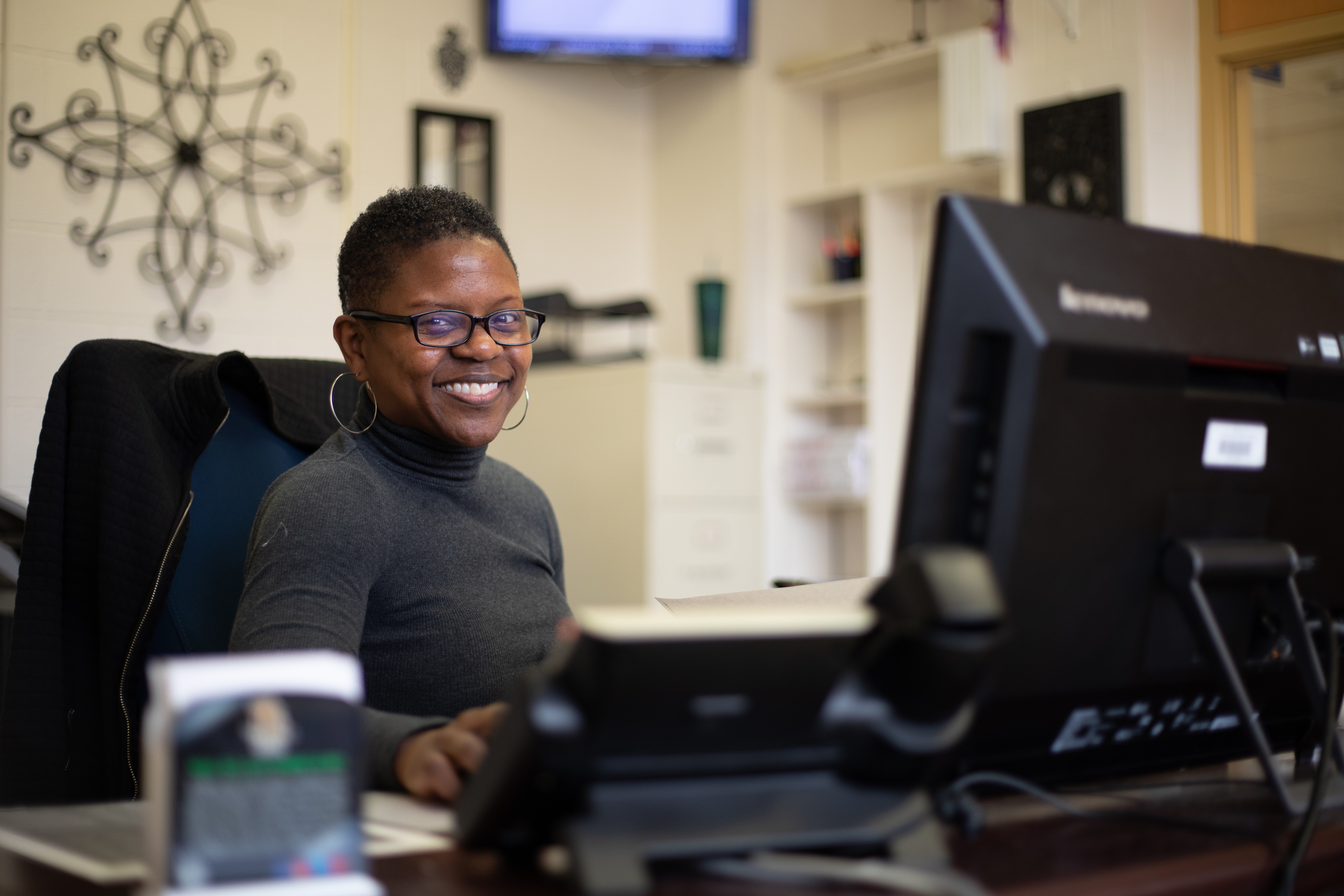 Image of African American wormen wearing glasses seated at a desk with a computer smiling at the camera