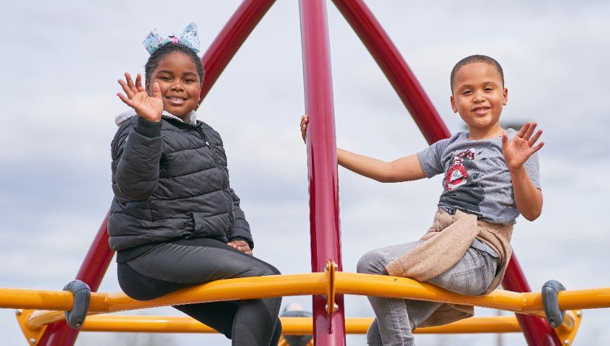 Two kids waving and sitting on playground equipment