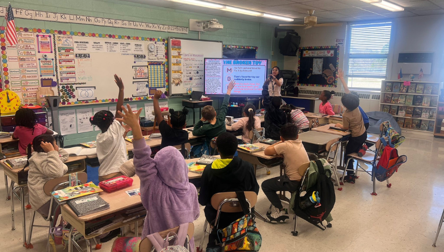 Elementary students raise their hands during a classroom lesson led by their teacher