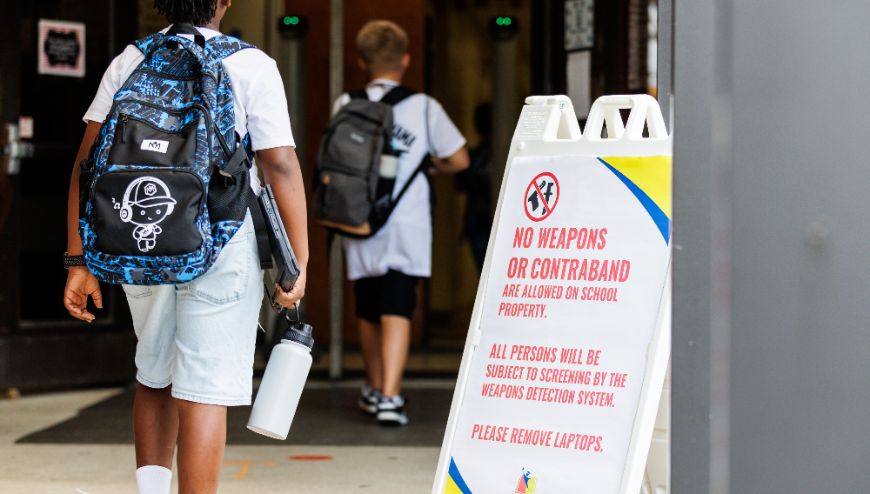 Image of student passing sign that says No weapons or contraband, are allowed on school property. All persons will be subject to screening by the eapons detection system. Please remove laptops.