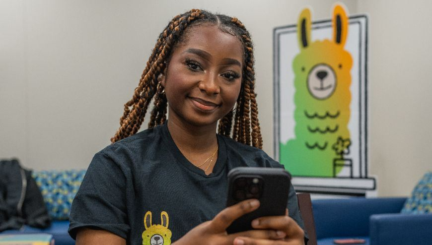 GIrl smiling holding a cell phone with an Alongside llama over her right shoulder.