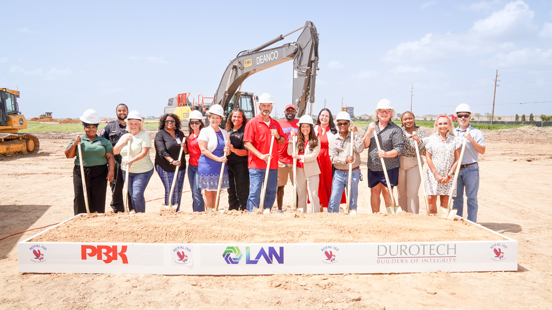 Group of Royal ISD leaders, staff, students, and partners wearing hard hats and holding shovels at the Royal High School groundbreaking site, with construction equipment behind them.