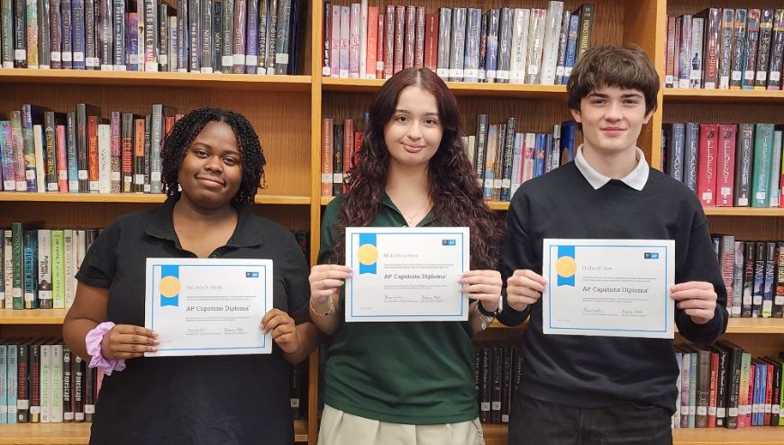 image of three students with their AP Capstone certificates with shelves of books behind thems