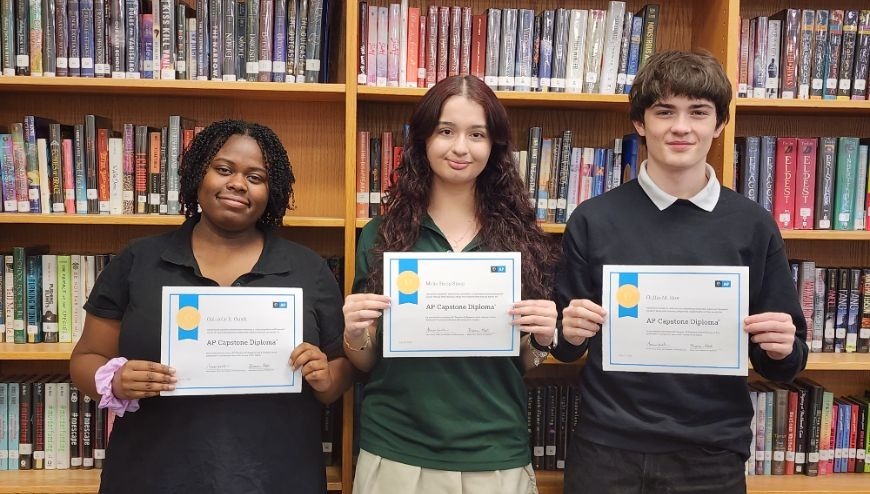 image of three students with their AP Capstone certificates with shelves of books behind thems