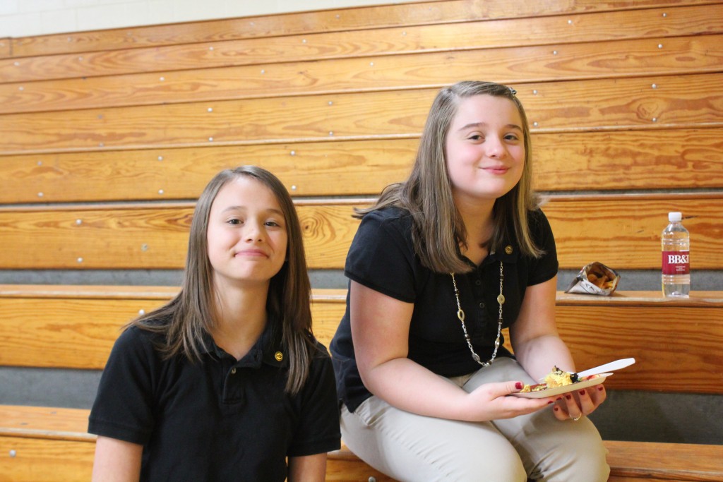 Two girls sitting on bleachers, chatting and laughing during a school basketball game.