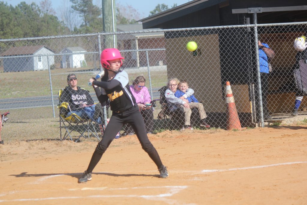 A girl in a pink helmet and black uniform swinging a bat.