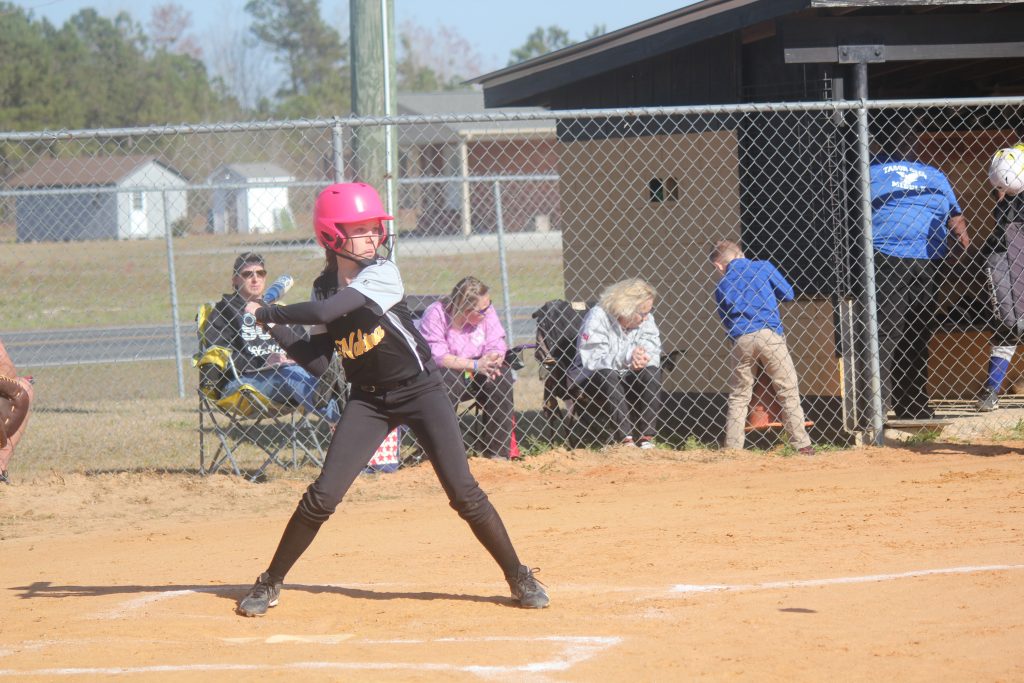 A girl in a pink helmet and black uniform swinging a bat.