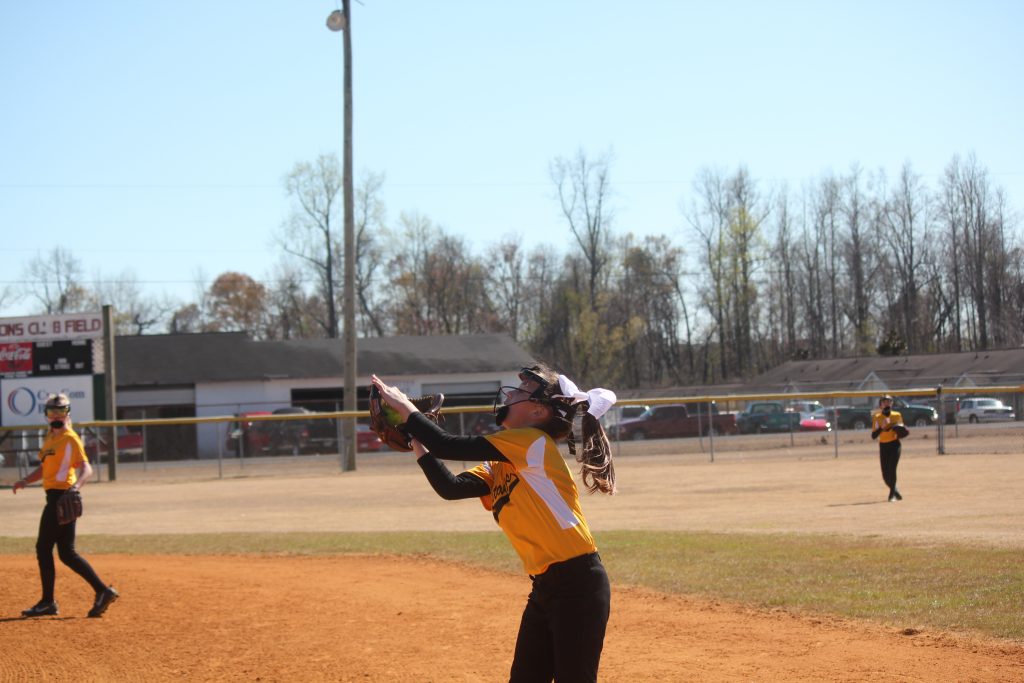 A girl in a yellow and black uniform throwing a ball on a sunny day.