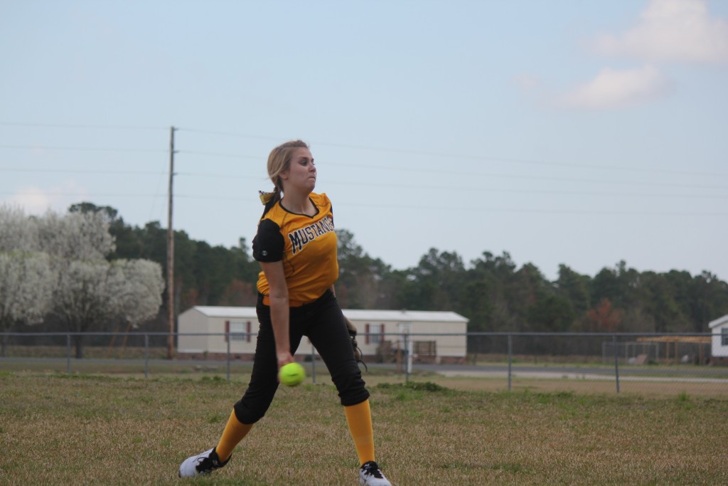 A girl in a yellow and black uniform pitching a ball on a sunny day.