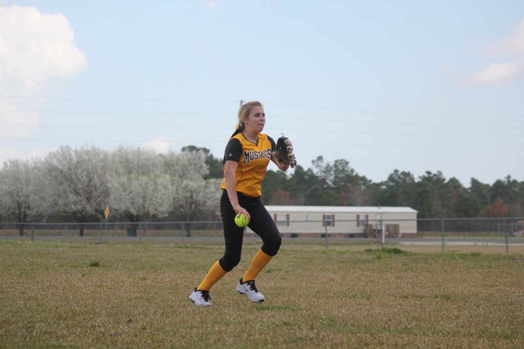 Young female athlete in yellow and black uniform pitching a softball on the field.