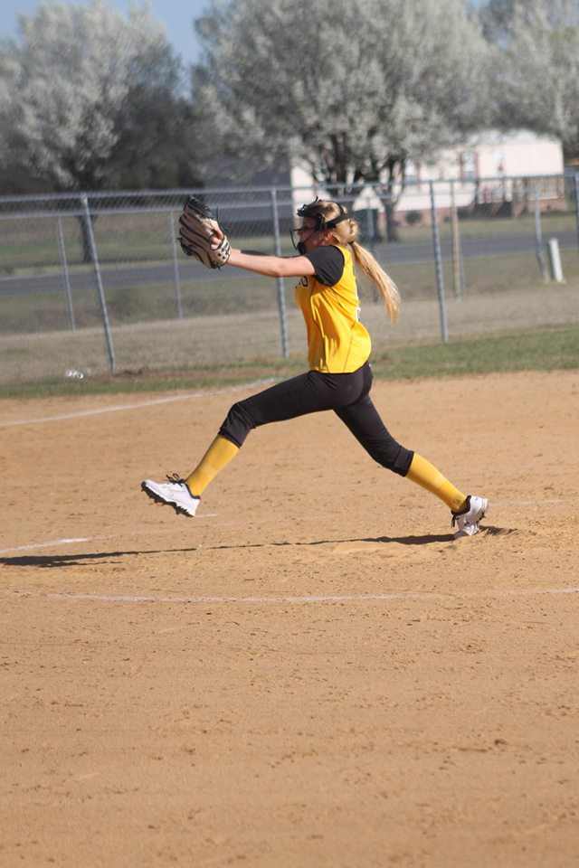 A young female athlete in a yellow and black uniform throwing a ball on a sports field.