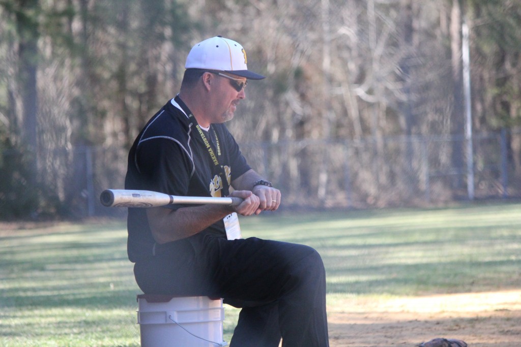 A man sitting on a bucket outdoors, holding a baseball bat.