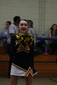 Cheerleader in black and yellow uniform performing energetic routine at a football game.