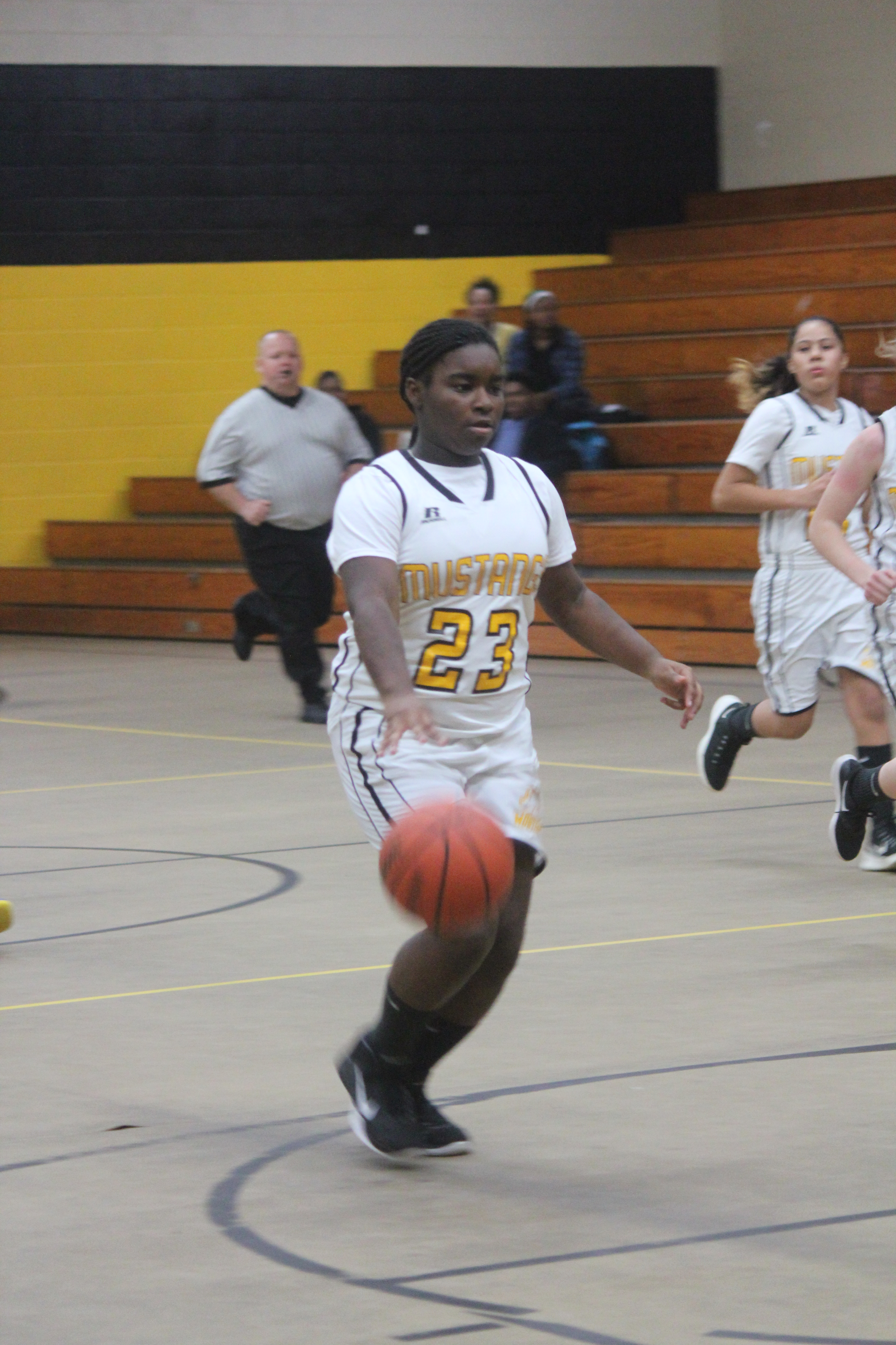 Young female athlete in white and yellow uniform dribbling basketball on court.