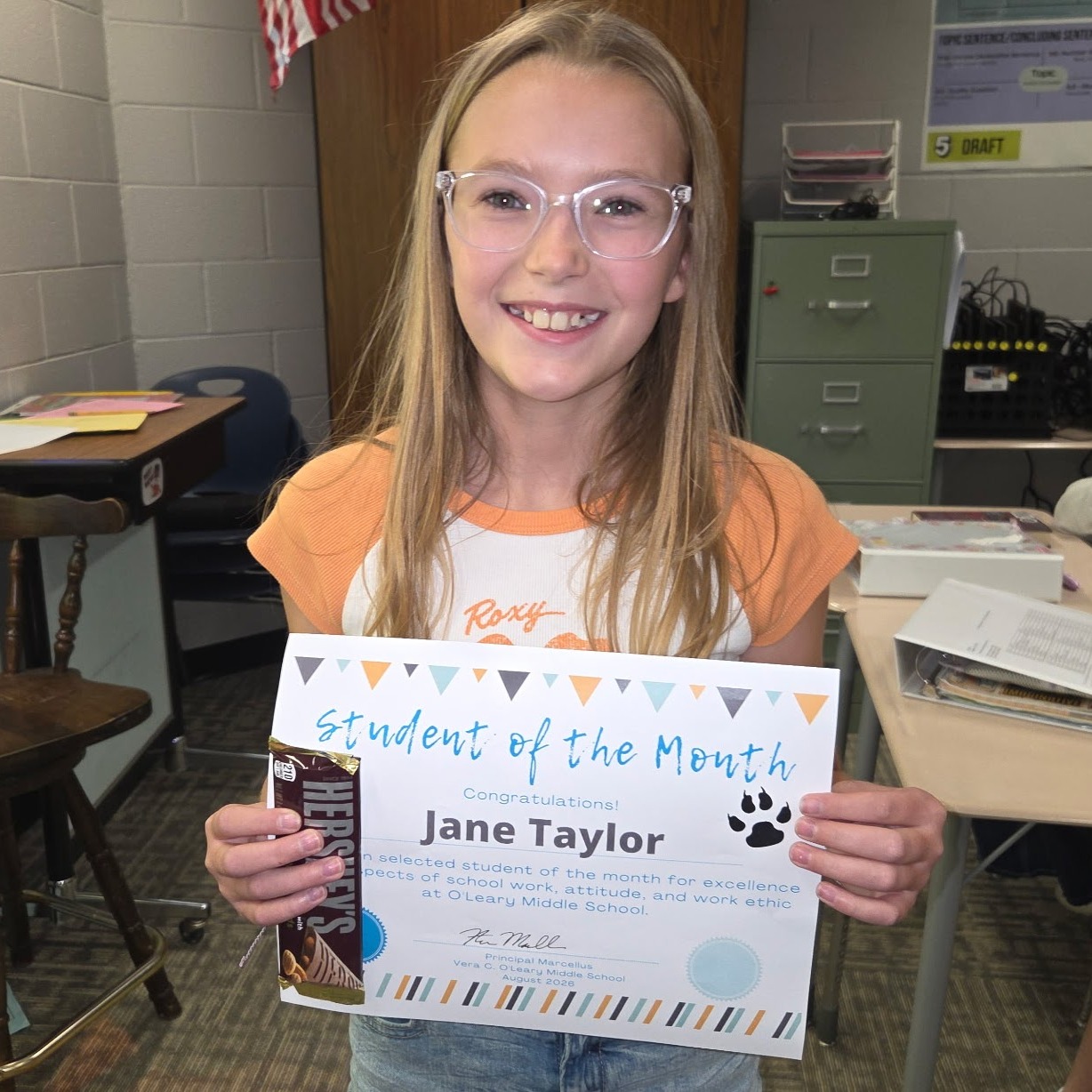 A young girl in a classroom holds a certificate, smiling and showing it to the camera.