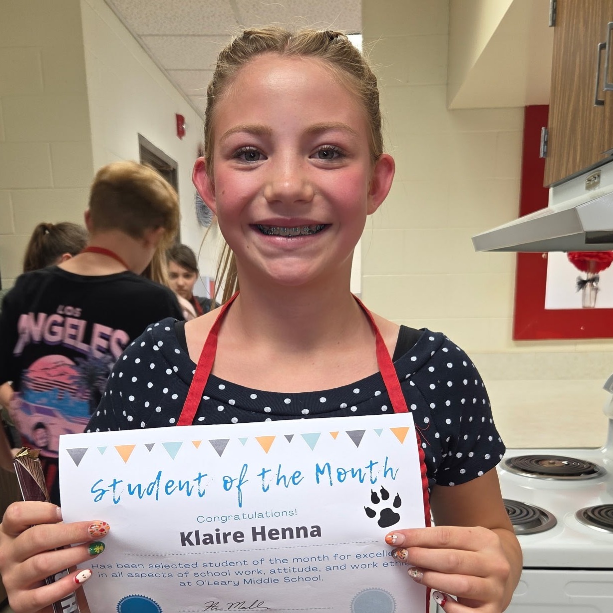 A girl holds a certificate labeled "Student of the Month" while wearing a red apron. Behind her, others are present in a kitchen.
