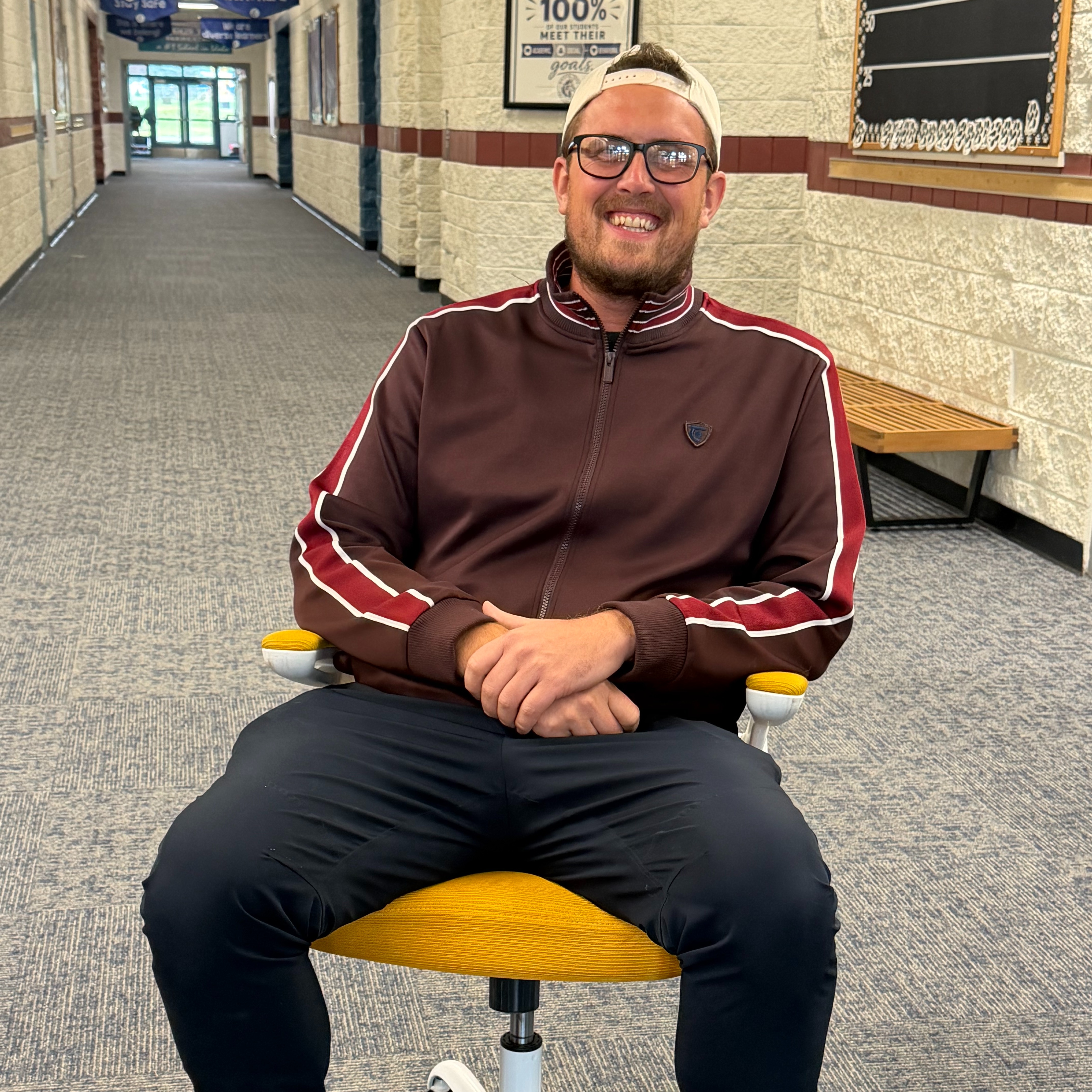 Person in a brown jacket, black pants, and glasses sits in a chair with hands on knees.