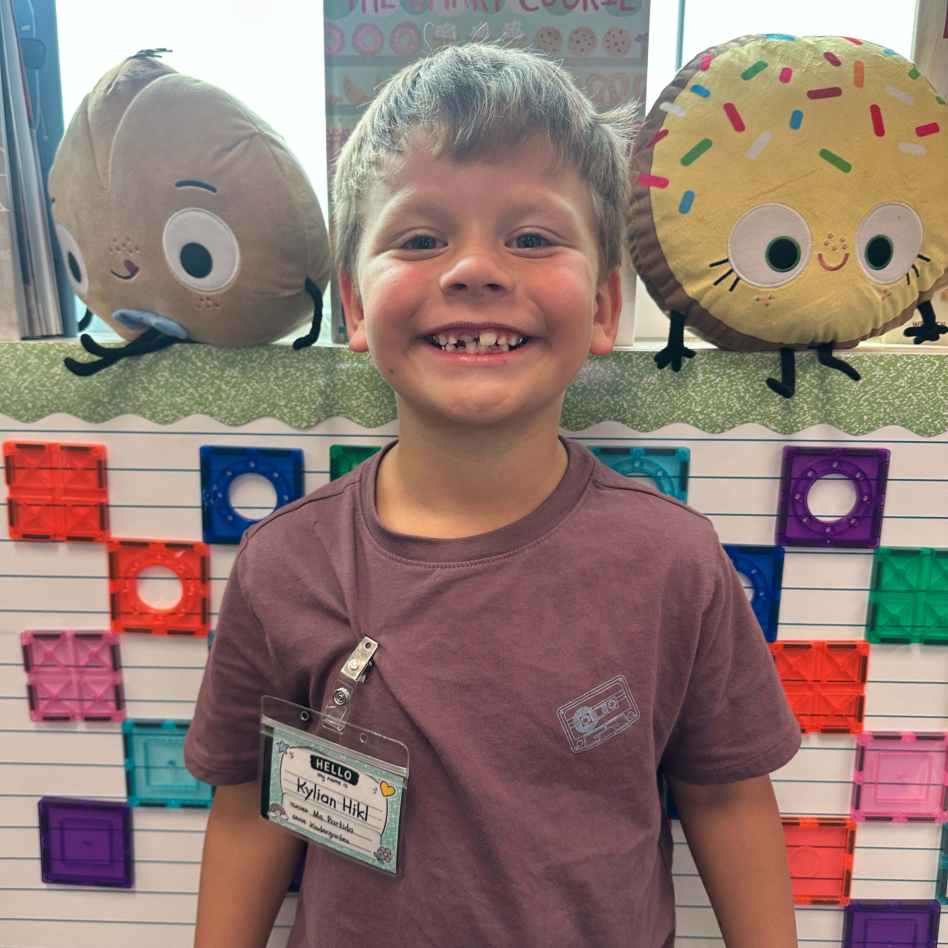 A smiling child in a brown t-shirt stands next to colorful blocks and two large stuffed toys.