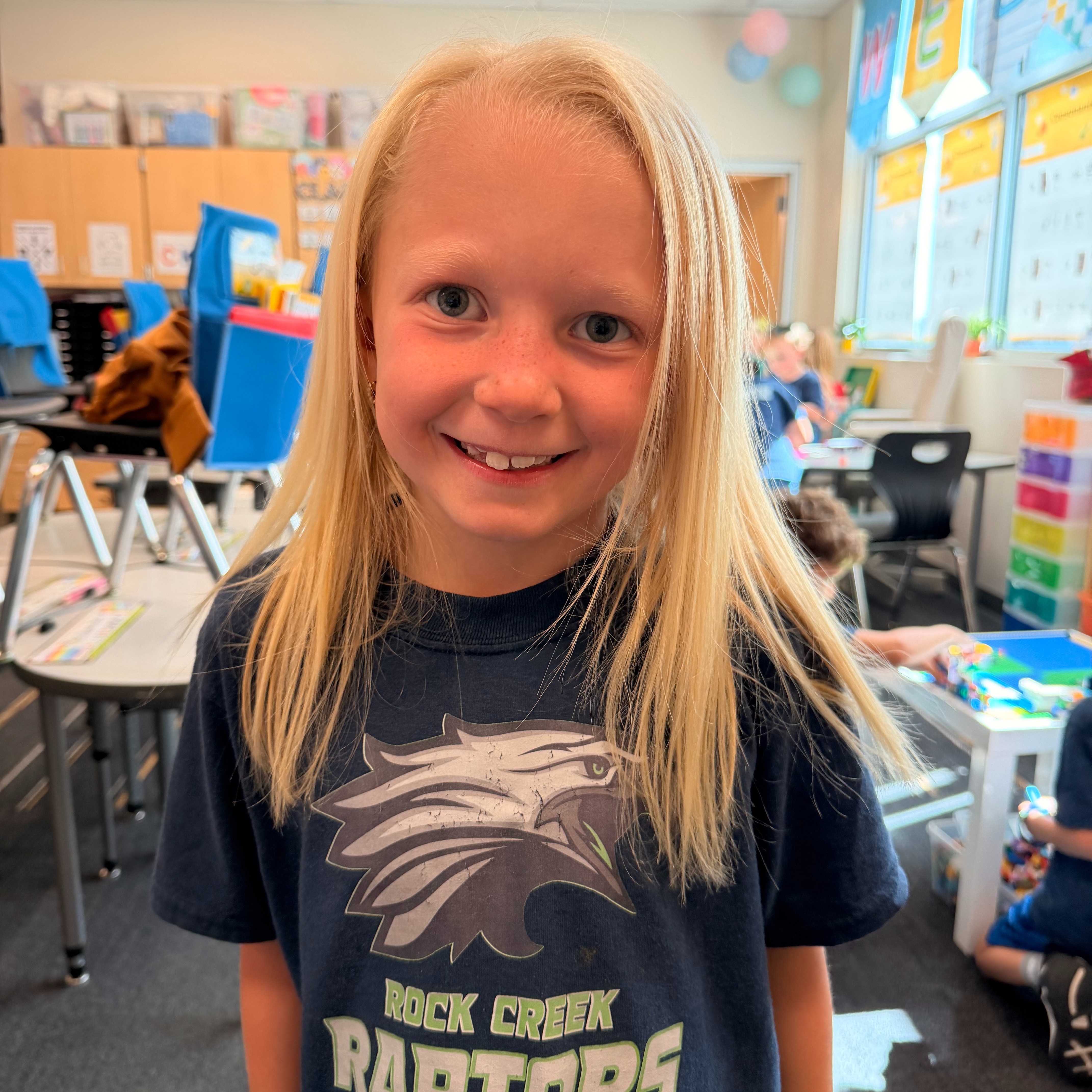 A young girl with blonde hair smiles in a classroom. She wears a blue T-shirt with "Rock Creek Raptors" printed on it.
