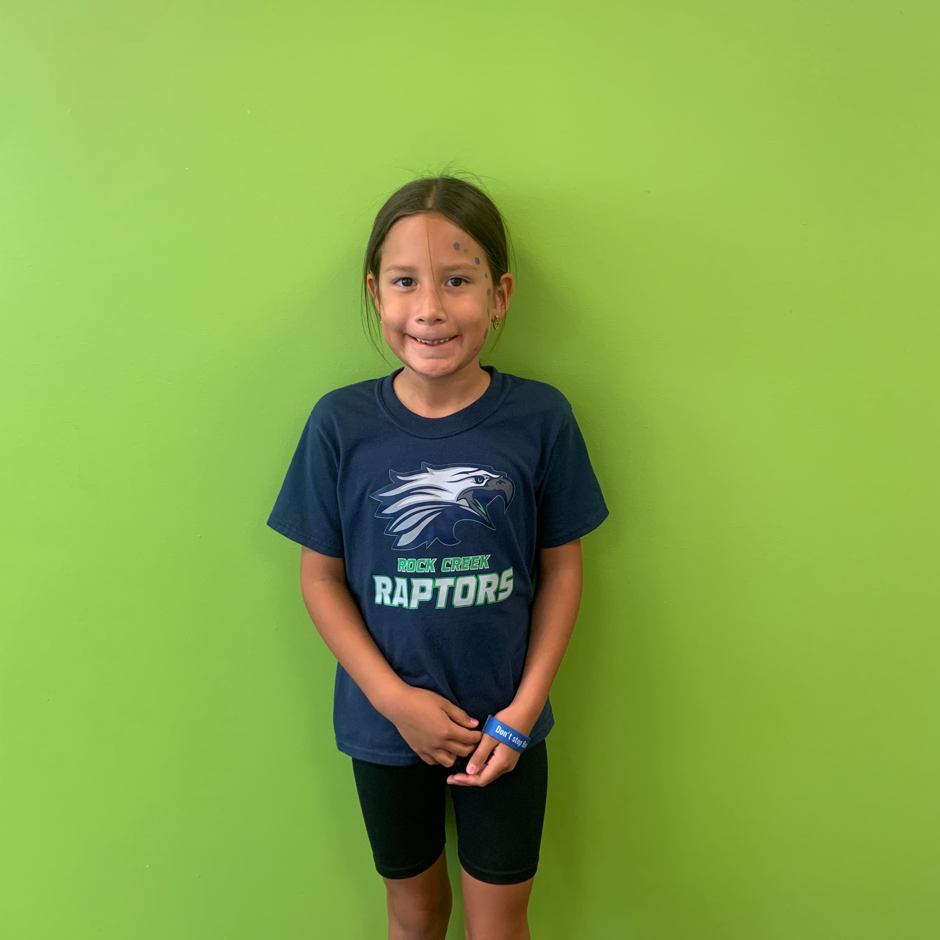 A young girl stands with a smile in front of a green wall, wearing a blue T-shirt.
