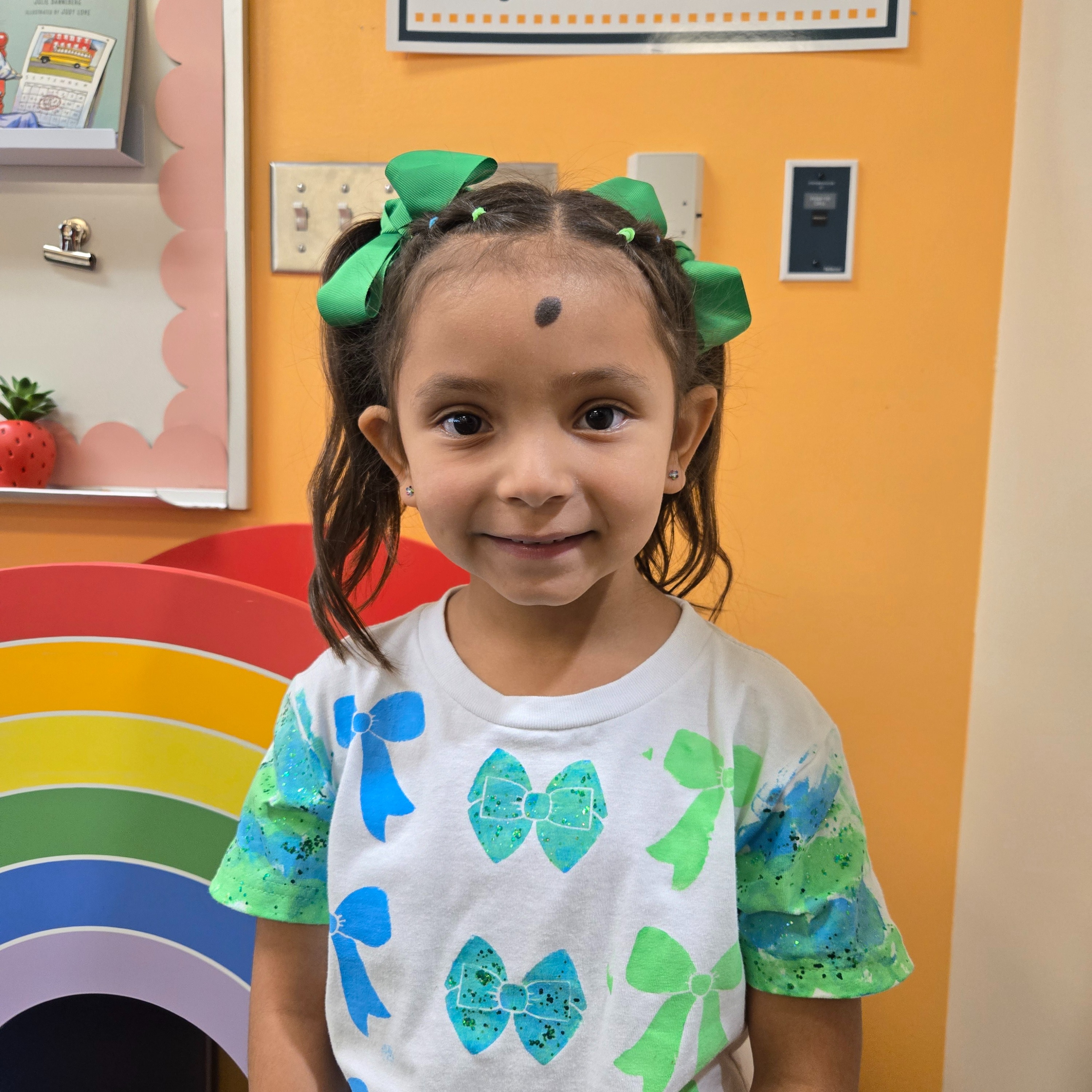 A young girl with green bows in her hair stands in a classroom. A colorful rainbow arch is behind her.