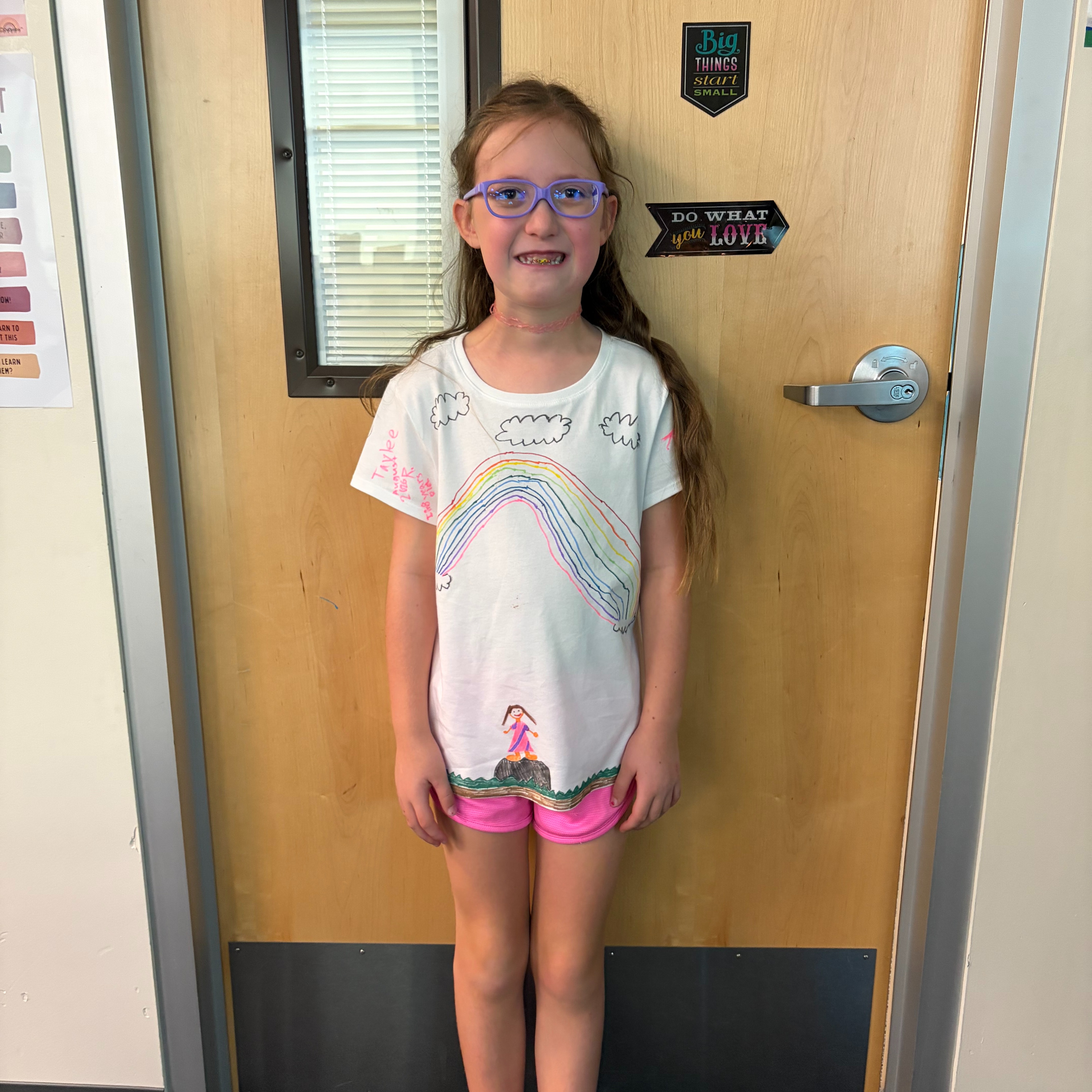 A young girl in glasses and sneakers stands in front of a wooden door with a window.