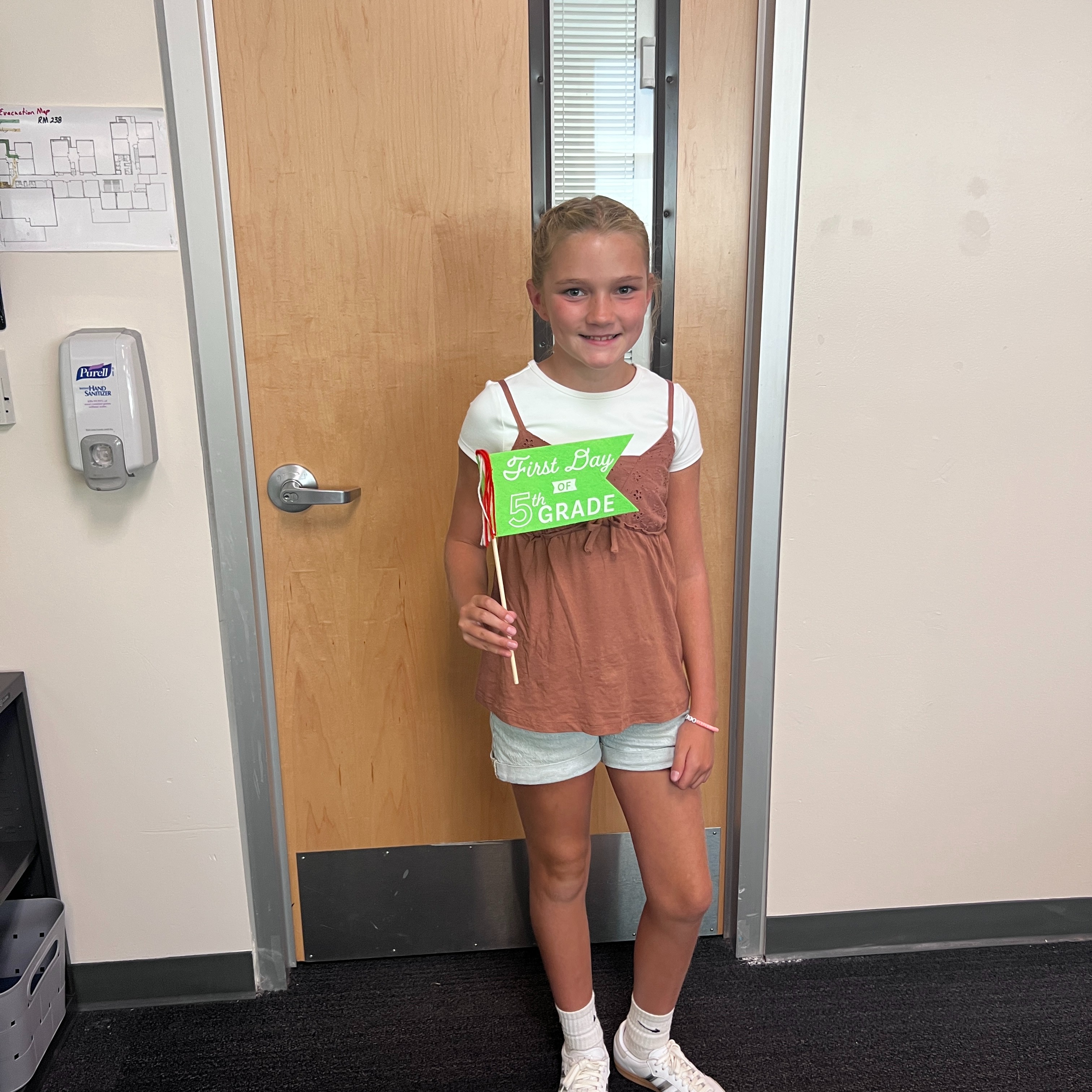 A young girl stands in a room, holding a green banner, and smiling. A wooden door is behind her.