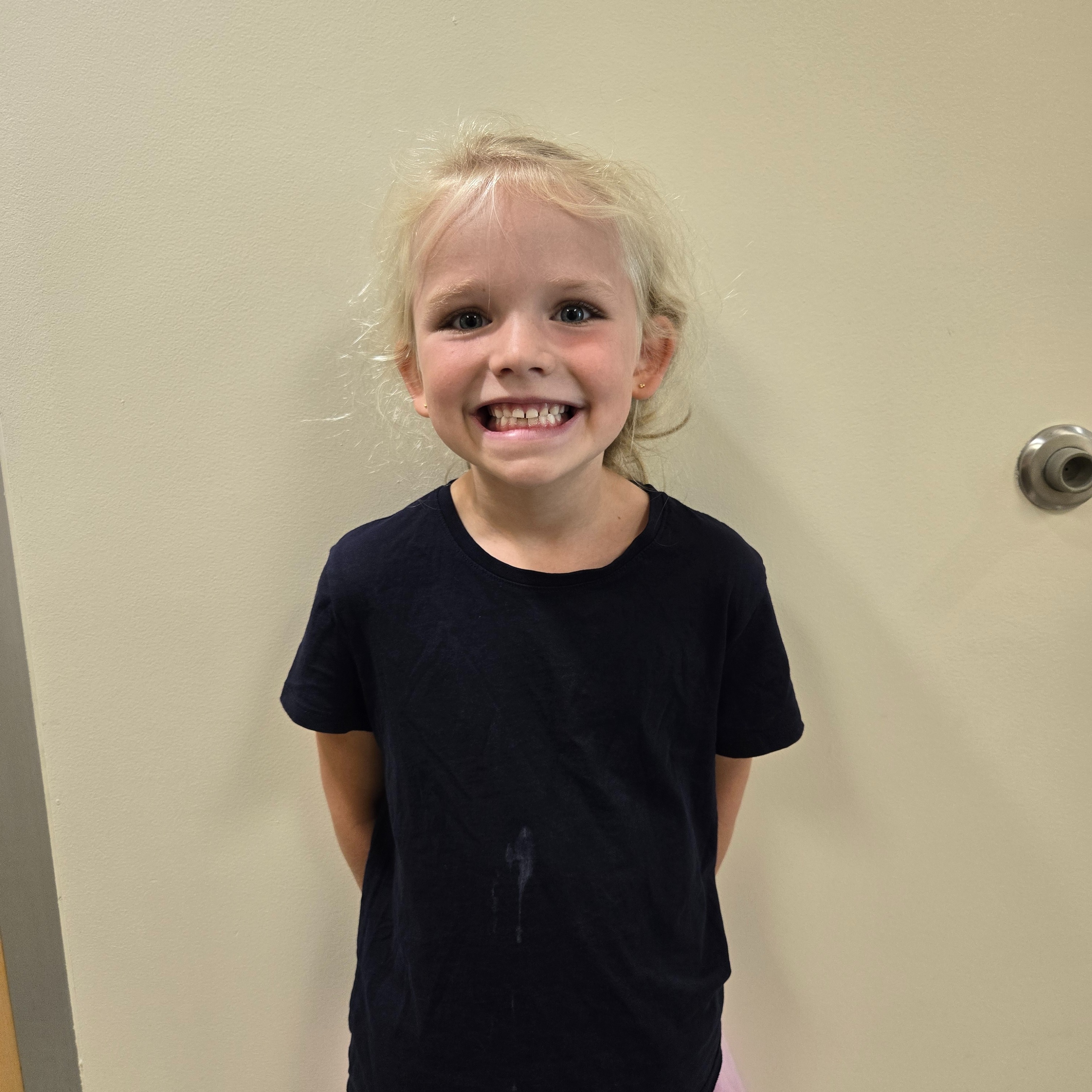 A girl with blond hair, wearing a black shirt and pink skirt, stands and smiles in front of a white wall.