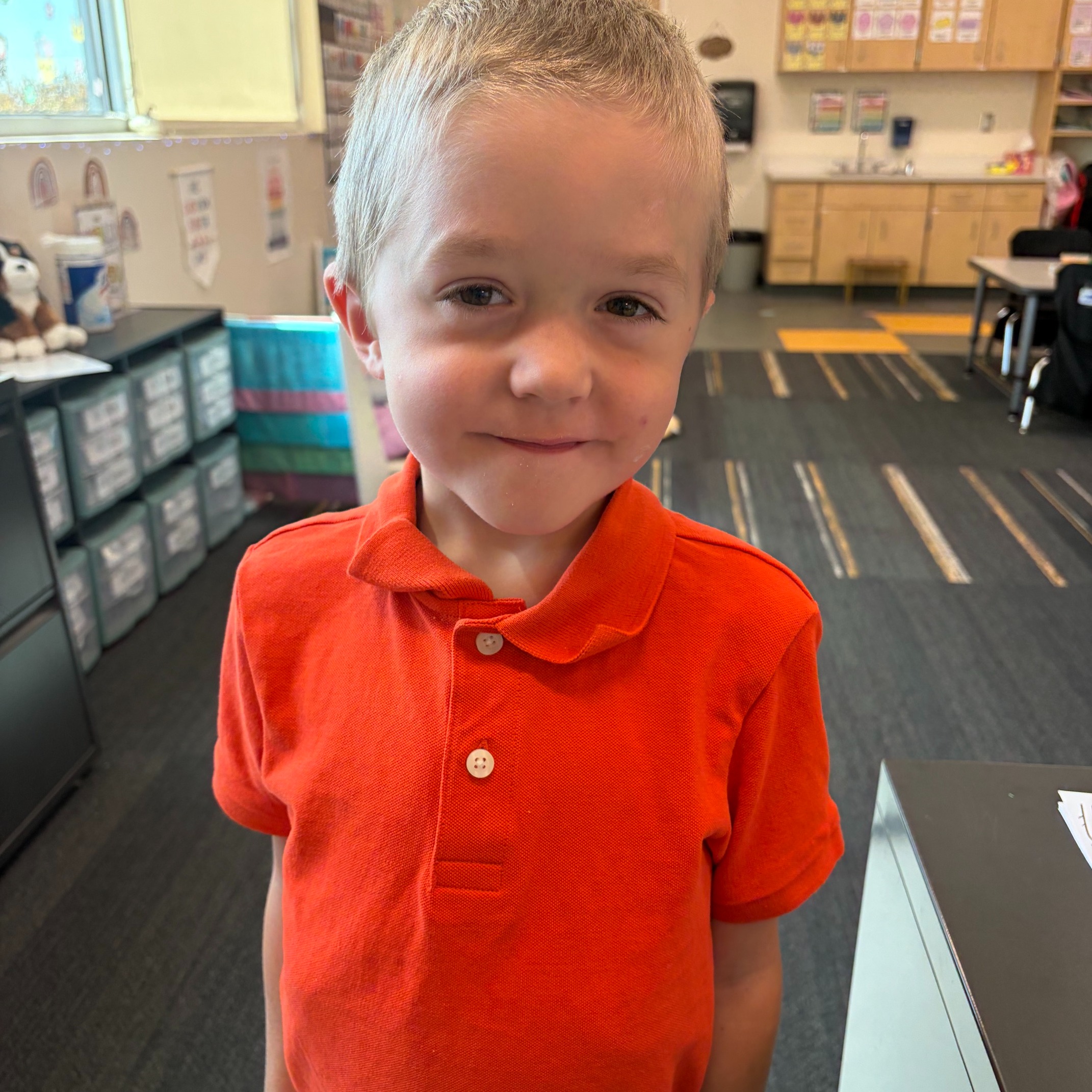 A young boy in an orange polo stands in a classroom with desks, storage cabinets, and a window.