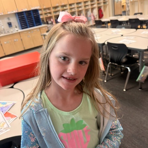 A young girl with blonde hair, smiling, in a classroom with desks and chairs in the background.