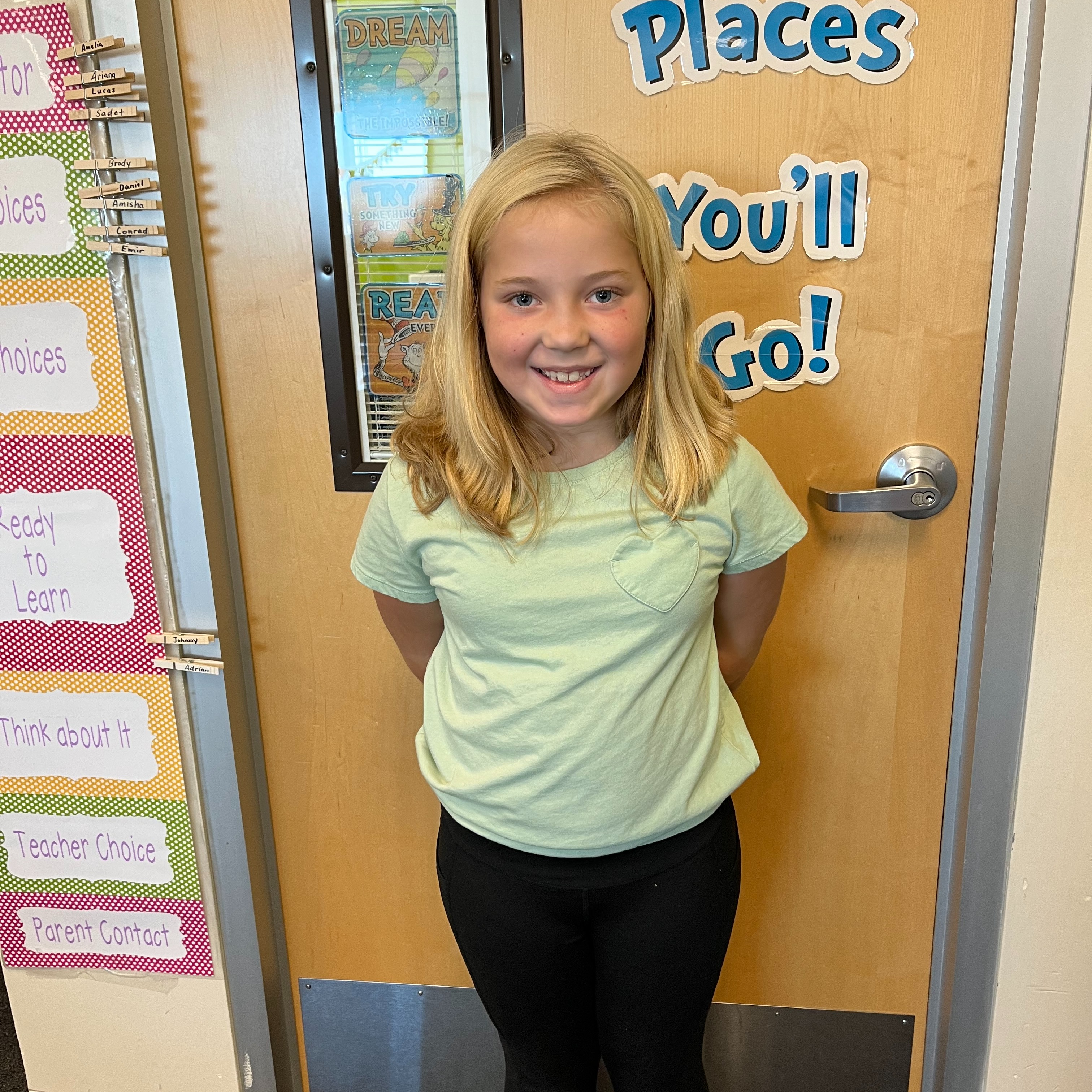 A young girl in a green shirt and black pants stands in front of a door. A bulletin board with colorful papers is on the left.