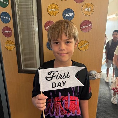 A young boy holds a white banner with "First Day" written on it in front of a wall with circles.