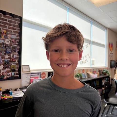 A boy smiles inside a classroom with a brick wall, a large window, and shelves behind him.