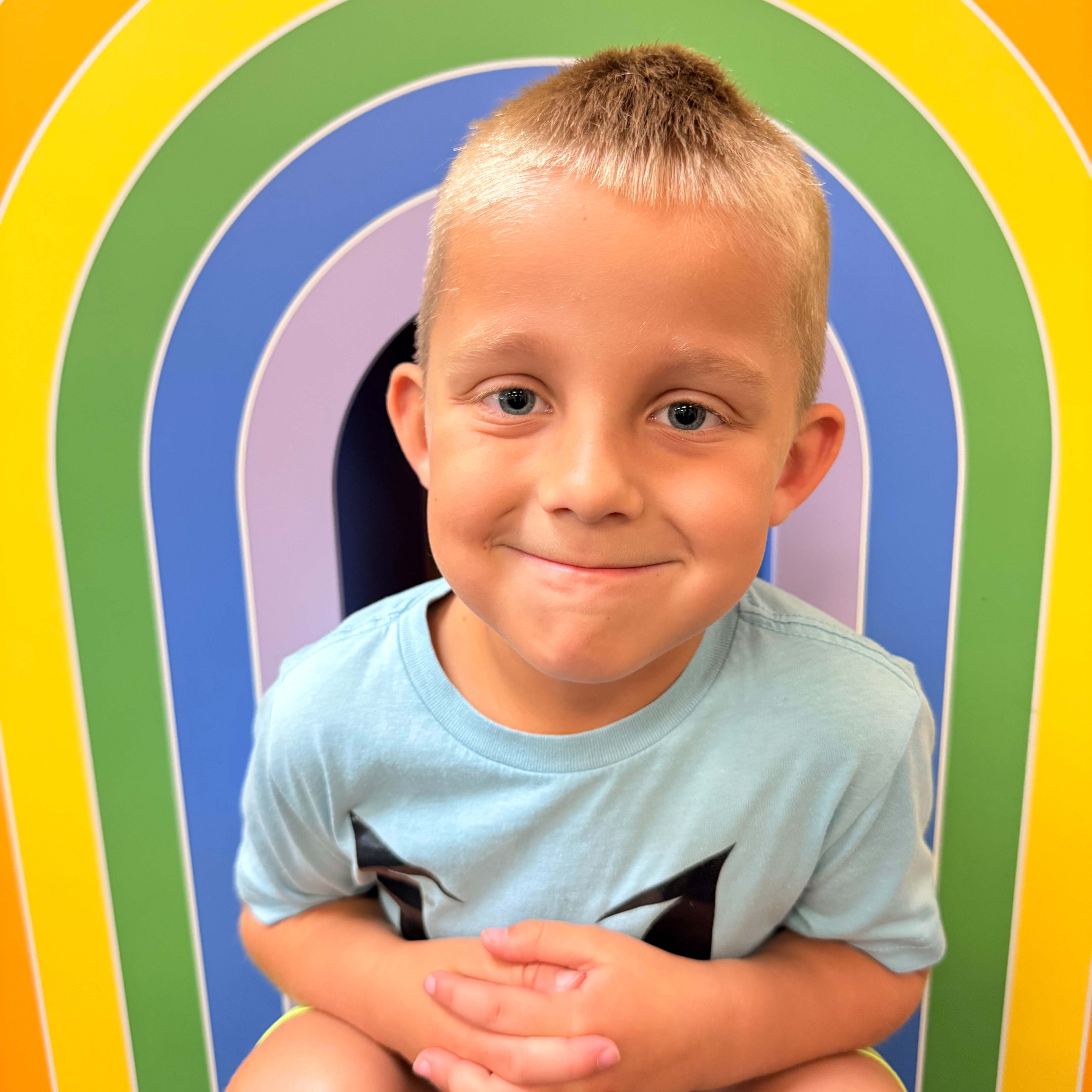 A young boy wearing a light blue t-shirt is sitting with a smile in front of a colorful rainbow backdrop.