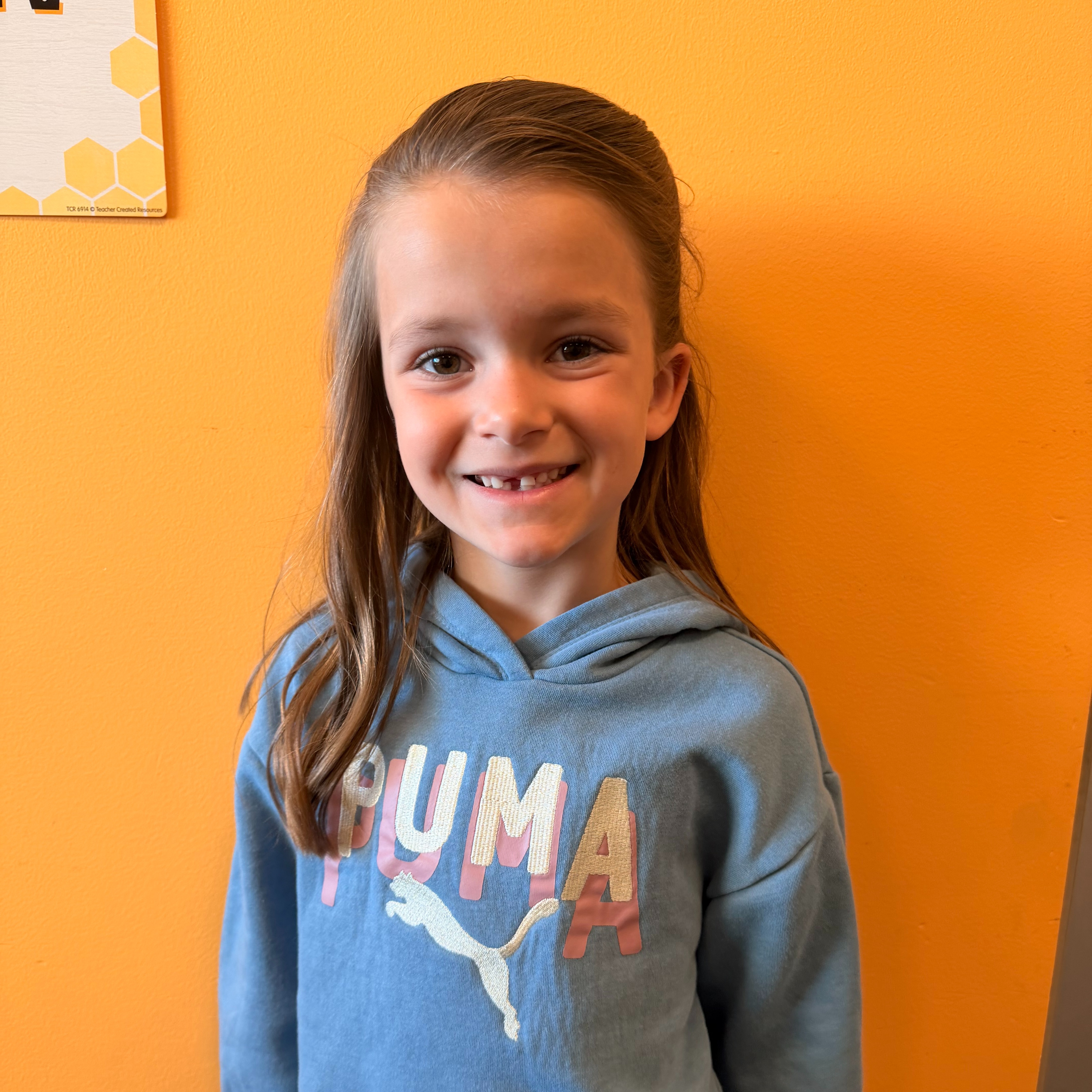 A girl stands before a yellow wall, wearing a blue hoodie with the word Puma. She smiles with her mouth open.