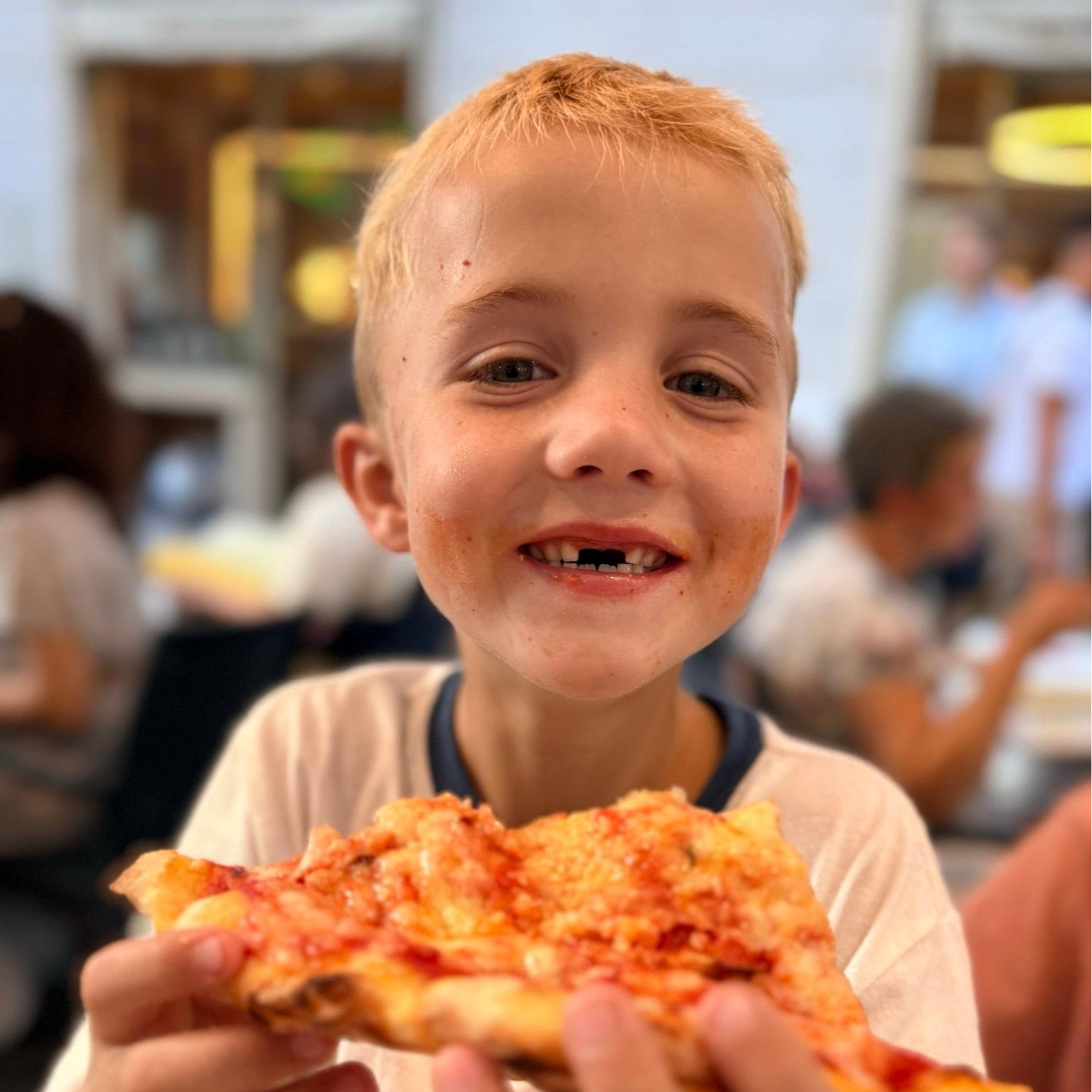 A child holds a slice of pizza, smiling and eating. Background includes other people seated at tables.