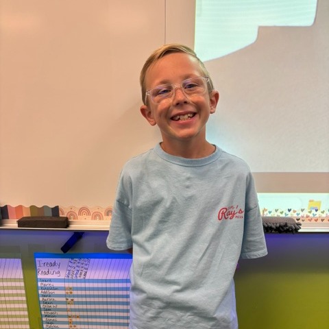 A boy in glasses and a blue t-shirt stands in front of a classroom wall, smiling.