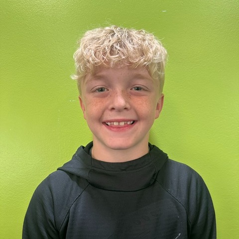 A boy with blonde hair smiles in front of a green wall, wearing a black hoodie.